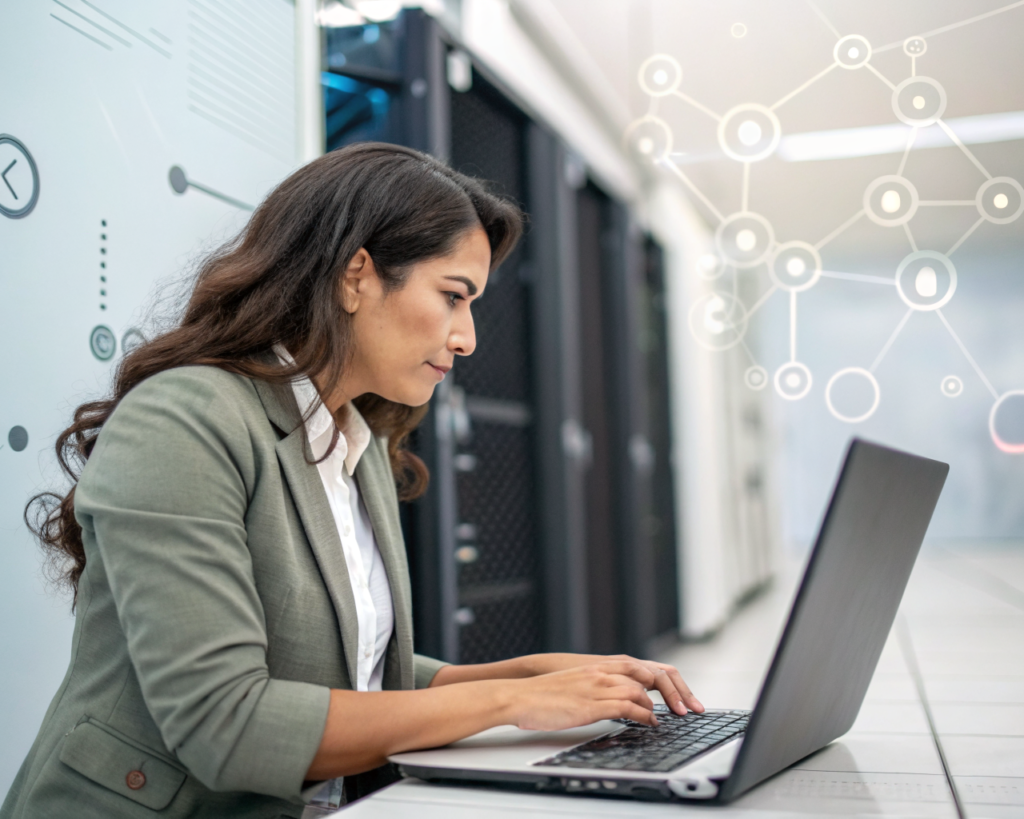 Mujer latinoamericana profesional, con expresión decidida y segura, trabaja en una laptop rodeada de equipo tecnológico y símbolos digitales de redes en un espacio moderno, con el teclado de la computadora y su rostro claramente enfocados y el fondo desenfocado.