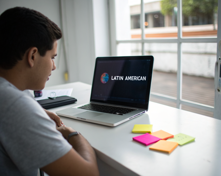 Persona latinoamericana genérica con ropa casual concentrada frente a un portátil moderno en un escritorio minimalista, rodeado de notas adhesivas de colores y una libreta cerrada. La luz natural de una ventana resalta la pantalla y manos, transmitiendo determinación y curiosidad, con fondo desenfocado y enfoque en las texturas del laptop y las notas escritas a mano.